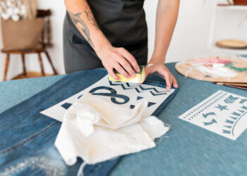 The image features a person customizing denim fabric using stencils and a sponge to apply paint. The individual, wearing a dark apron, presses the sponge over a stencil with bold designs, including chevrons and stars. Nearby, there are other stencils and tools like brushes, fabric scraps, and a palette with mixed paints, indicating an artistic workspace. The setting has a casual, creative vibe, with a focus on DIY fashion or fabric decoration. The background includes homey elements like a wooden chair and decor, enhancing the cozy and personalized atmosphere of the scene.