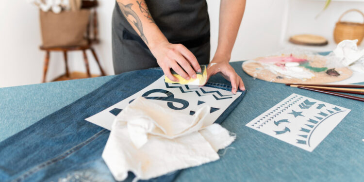 The image features a person customizing denim fabric using stencils and a sponge to apply paint. The individual, wearing a dark apron, presses the sponge over a stencil with bold designs, including chevrons and stars. Nearby, there are other stencils and tools like brushes, fabric scraps, and a palette with mixed paints, indicating an artistic workspace. The setting has a casual, creative vibe, with a focus on DIY fashion or fabric decoration. The background includes homey elements like a wooden chair and decor, enhancing the cozy and personalized atmosphere of the scene.